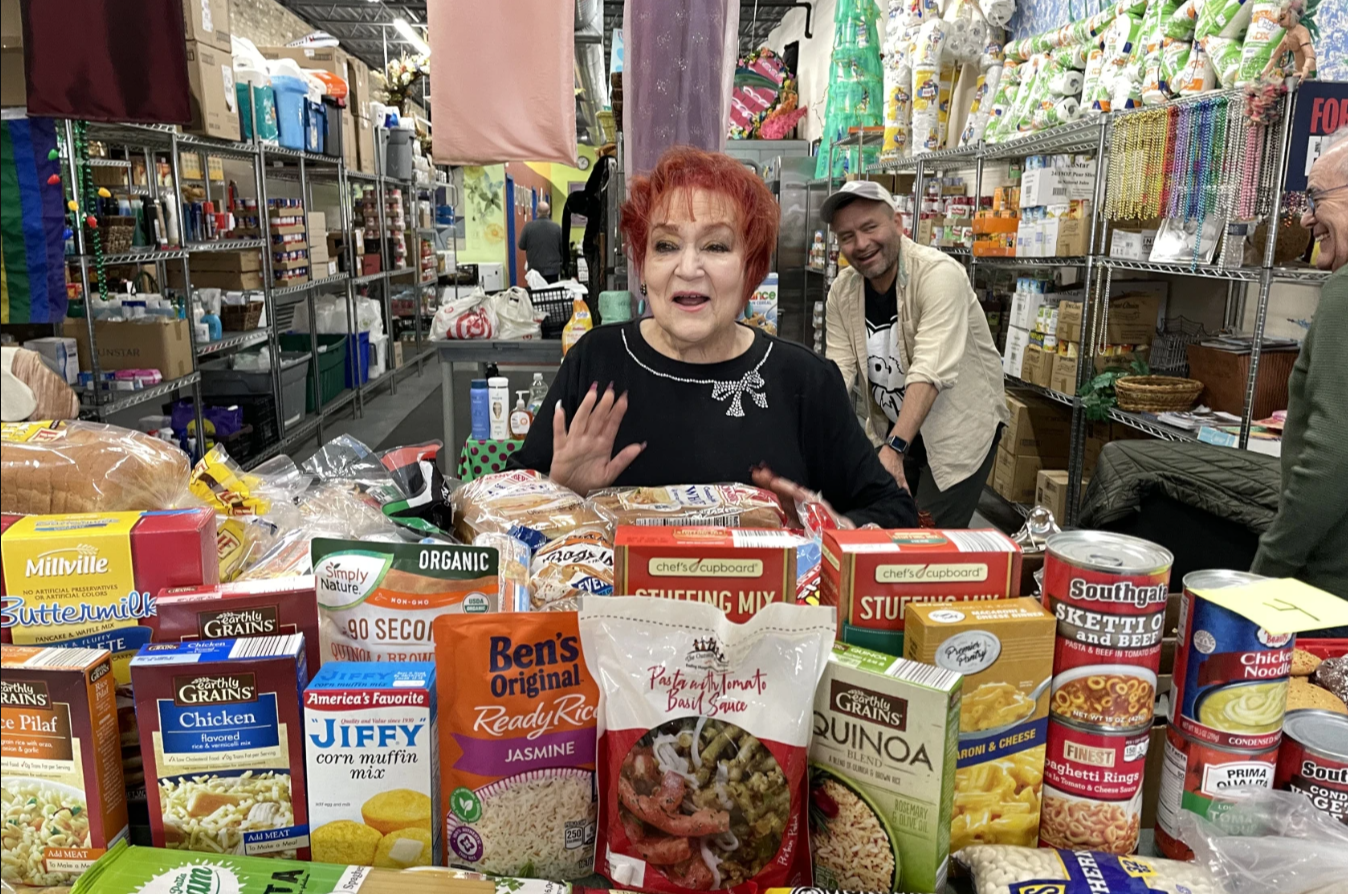 A photo of a red haired woman, Lori Cannon, standing behind a table full of shelf stable groceries. The woman is Lori Cannon.