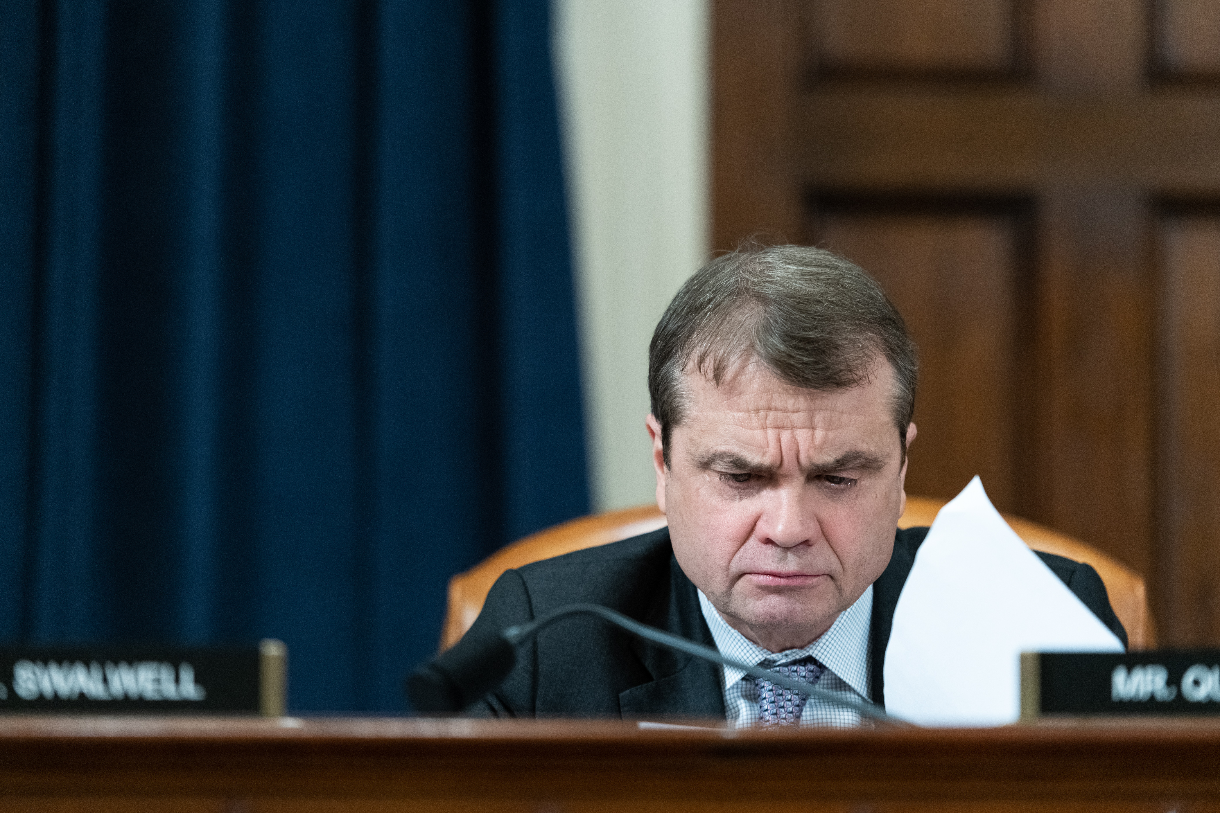 A man in a suit sits on a dais looking seriously at some papers. The man is Mike Quigley.