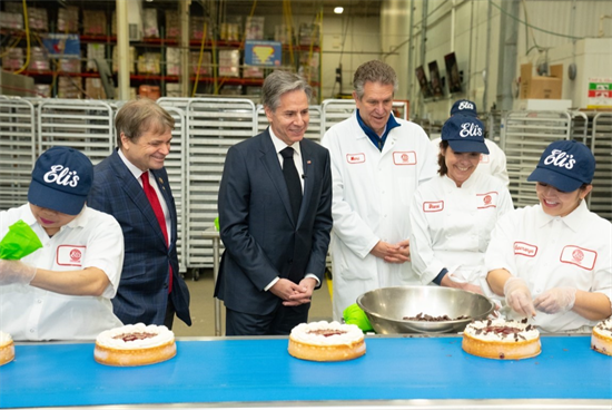 Quigley and Secretary Blinken smiling over a conveyer belt of Eli's cheesecakes